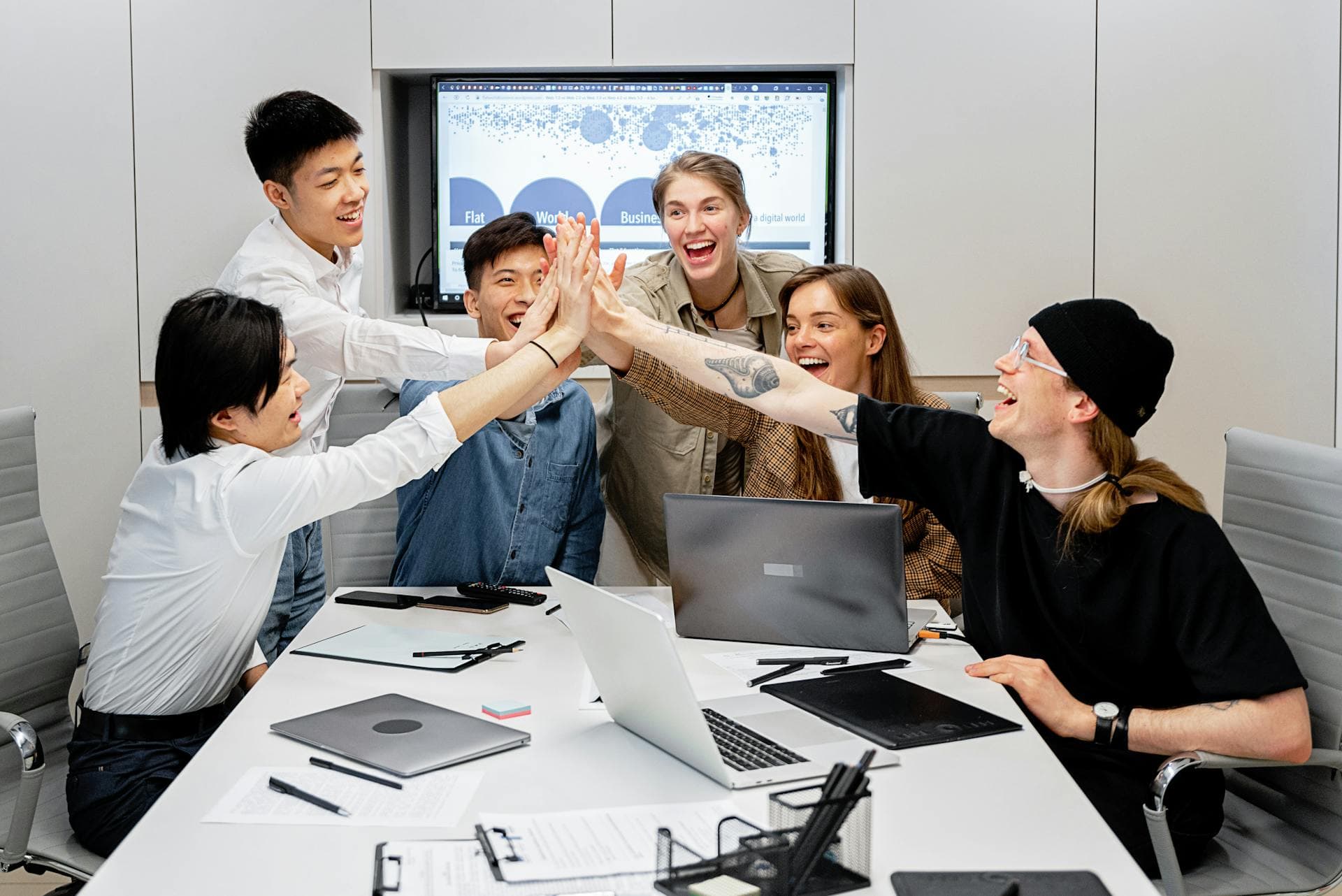 A marketing team celebrating around a conference table with laptops and a data dashboard in the background. The payoff when your GEO playbook starts landing you in AI recommendations.